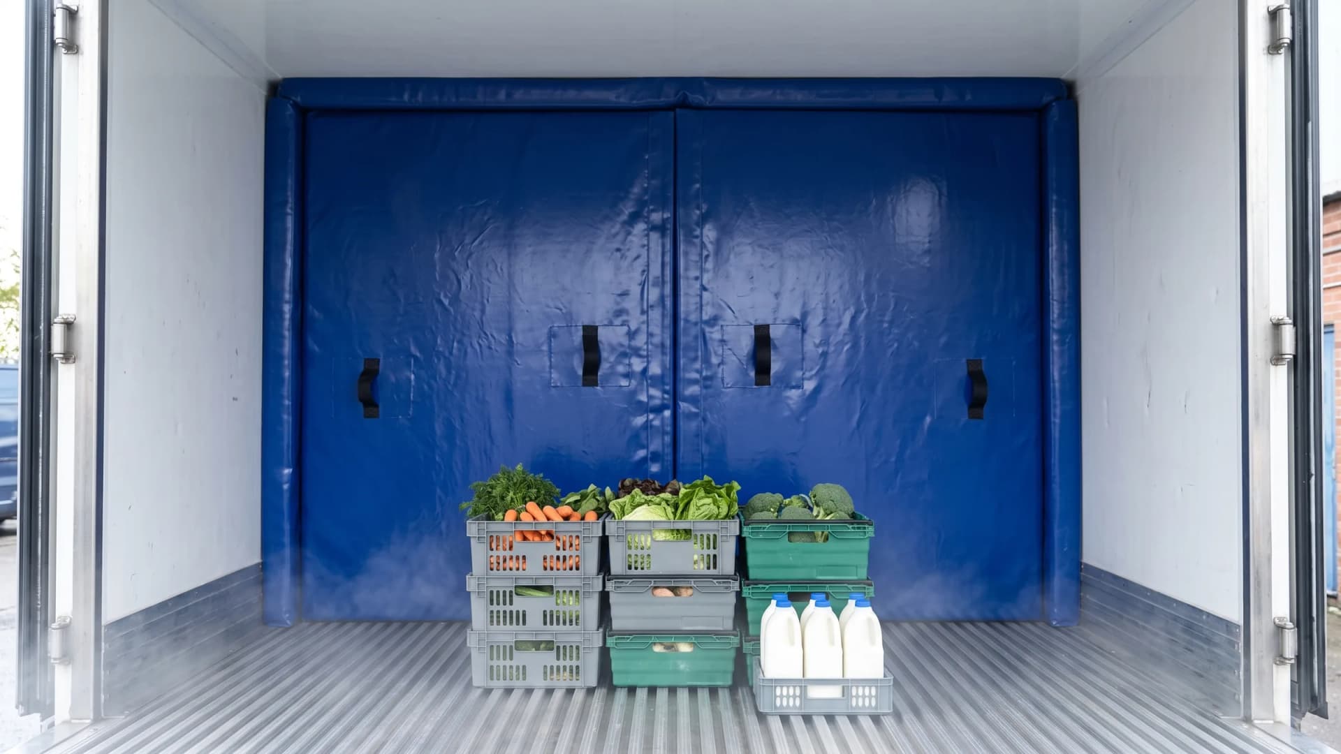 Royal-blue padded PVC bun partition filling the full width and height of a white refrigerated truck body, black webbing handles on its face, crates of fresh vegetables and milk bottles on the aluminium floor in front of it