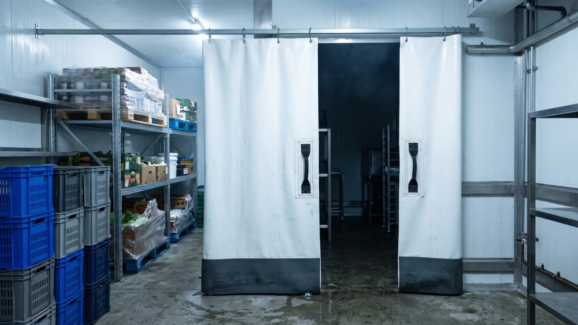 Two white insulated PVC thermal partition panels hung from a ceiling rail inside a walk-in cold room, each with a dark grey scuff board along its bottom edge and a black reinforced webbing handle stitched to its face, chilled product on racking to one side and a darker zone visible through the gap between them